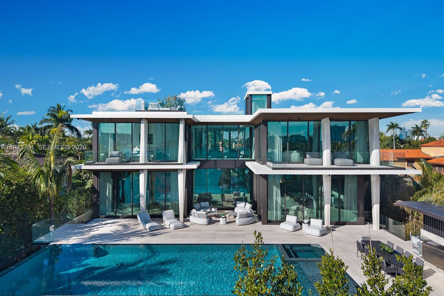 This modern, multi-level luxury residence features a striking contemporary design with extensive floor-to-ceiling glass walls that blur the lines between indoor and outdoor living. The rear exterior showcases a spacious stone patio furnished with lounge seating, leading directly to a pristine, rectangular swimming pool. The architectural aesthetic is defined by clean lines, white structural accents, and a sophisticated, open-plan layout that captures abundant natural light.