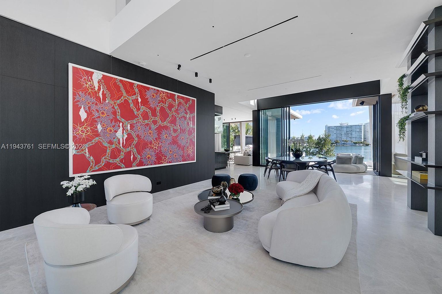 This expansive, modern living room features a striking large-scale red abstract art piece mounted on a dark accent wall, contrasting with the light-toned flooring and curved white furniture. The space flows seamlessly toward a wall of floor-to-ceiling glass doors that open to a waterfront view, creating an airy and sophisticated atmosphere. The open-concept layout includes a dining area in the background, emphasizing a luxurious, minimalist aesthetic perfect for high-end entertaining.