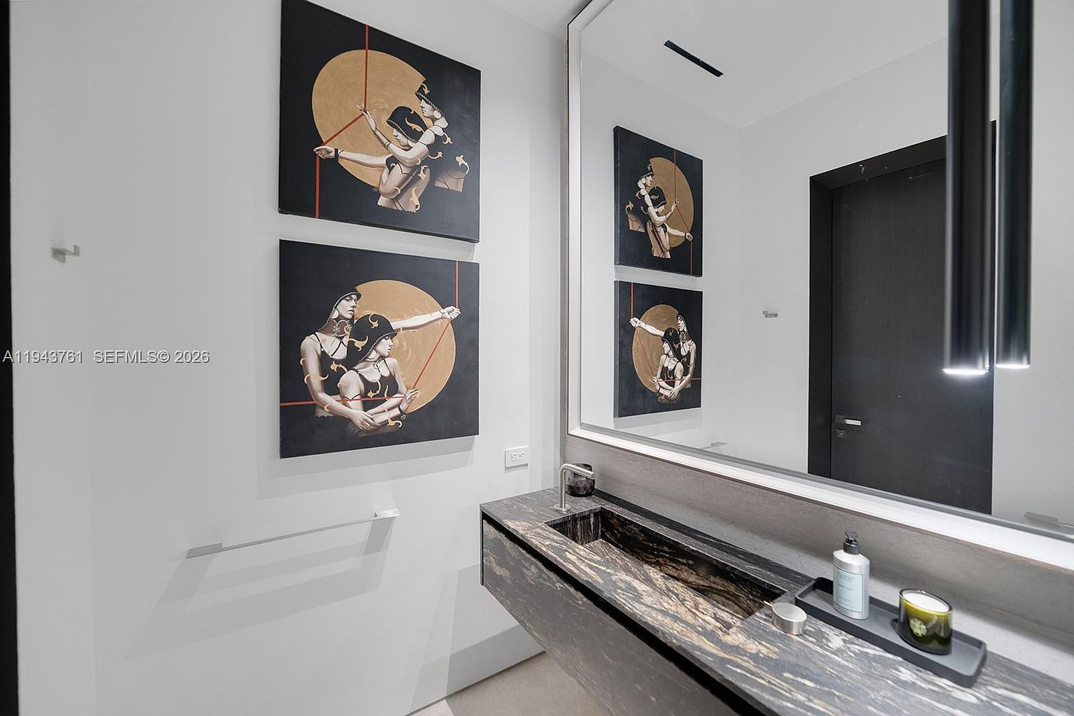 This modern powder room features a striking floating vanity with a dark, heavily veined stone countertop and integrated sink. The walls are adorned with two large, matching pieces of contemporary art, while a massive, frameless mirror reflects the space, enhancing the sense of depth. The minimalist aesthetic is complemented by a sleek wall-mounted faucet and a dark door, creating a sophisticated and high-end atmosphere.