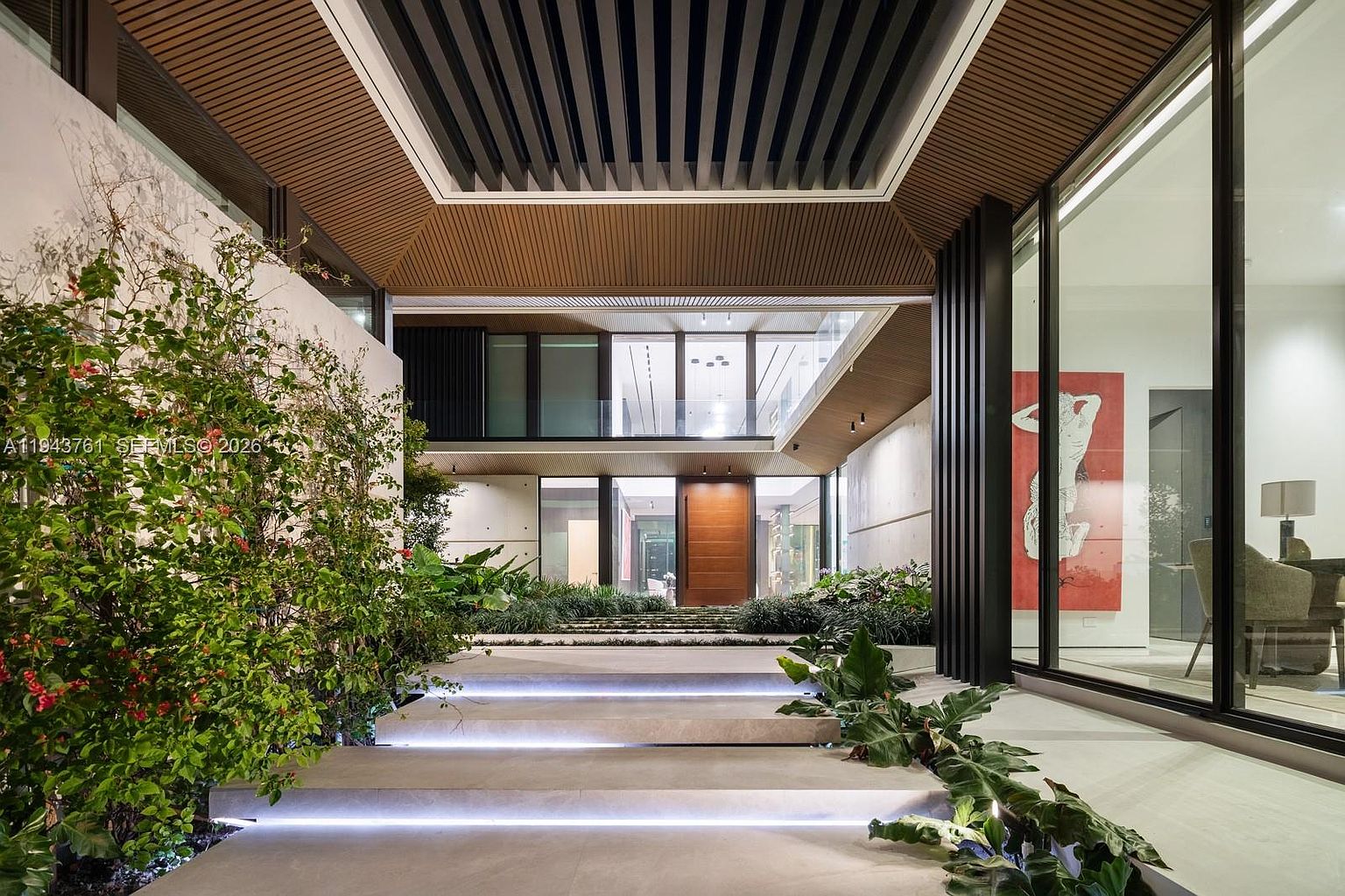 This striking modern entryway features a grand, tiered staircase illuminated by integrated LED lighting, leading up to a prominent wooden front door. The space is framed by a dramatic slatted wood ceiling and lush, integrated landscaping that softens the clean lines of the concrete and glass architecture. The perspective is a wide-angle, eye-level shot that emphasizes the depth and luxurious, resort-like atmosphere of the home's entrance.