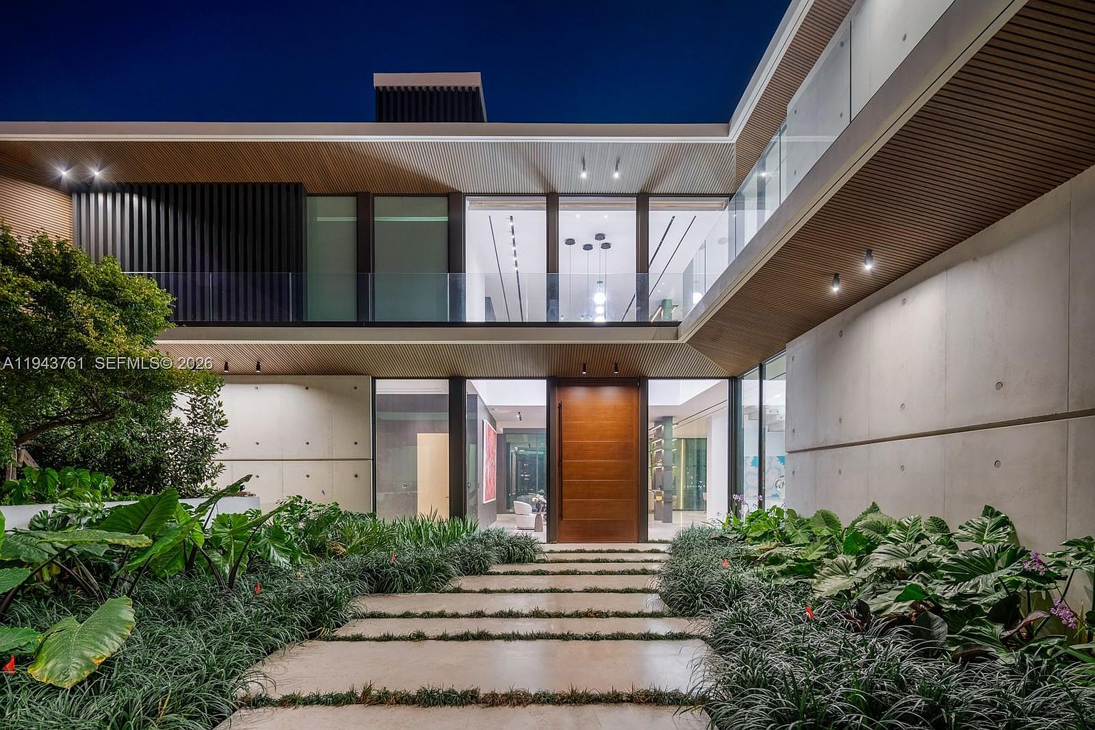 This striking modern residence features a grand entryway defined by a massive, warm-toned wooden door set against a backdrop of smooth concrete walls and expansive glass panels. A symmetrical stone pathway leads through a lush, manicured garden with vibrant tropical foliage, creating a sophisticated and inviting approach. The upper level showcases a sleek balcony with glass railings, emphasizing the clean lines and contemporary architectural aesthetic of the home.