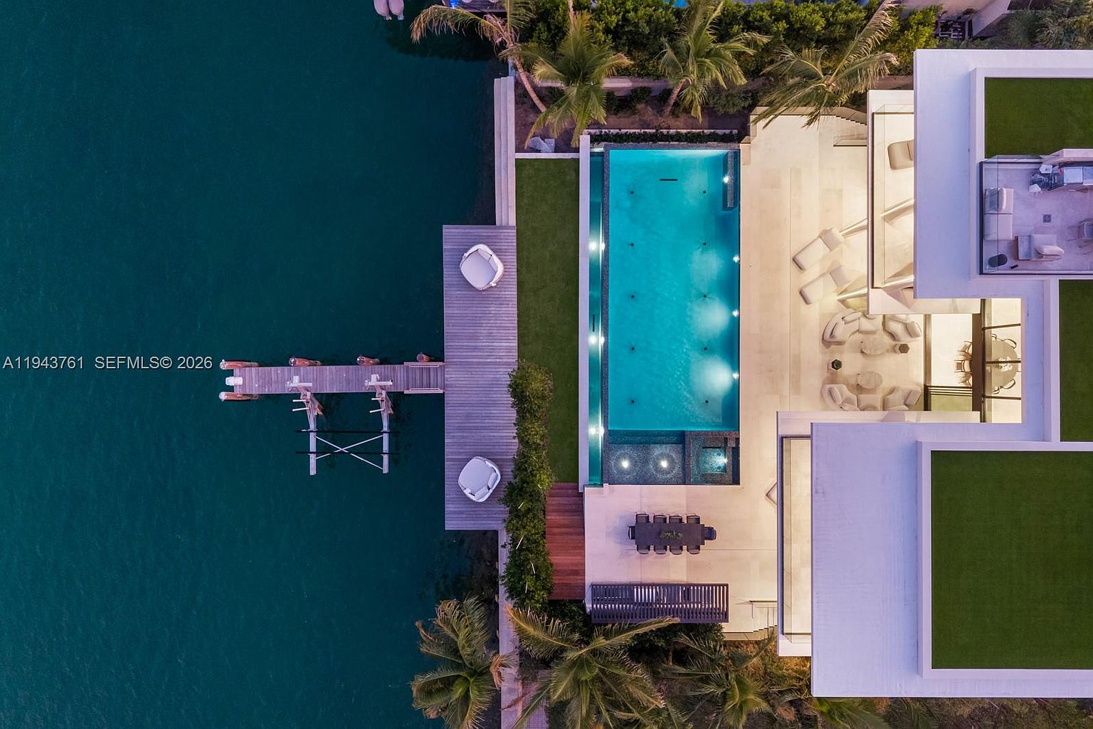 This high-angle aerial view captures a luxurious waterfront property featuring a long, rectangular swimming pool and an adjacent spa. The outdoor space includes a wooden deck with lounge seating, a dining area, and a private dock extending into the calm water. The modern architecture is complemented by lush palm trees and manicured green spaces, creating a serene and upscale atmosphere.