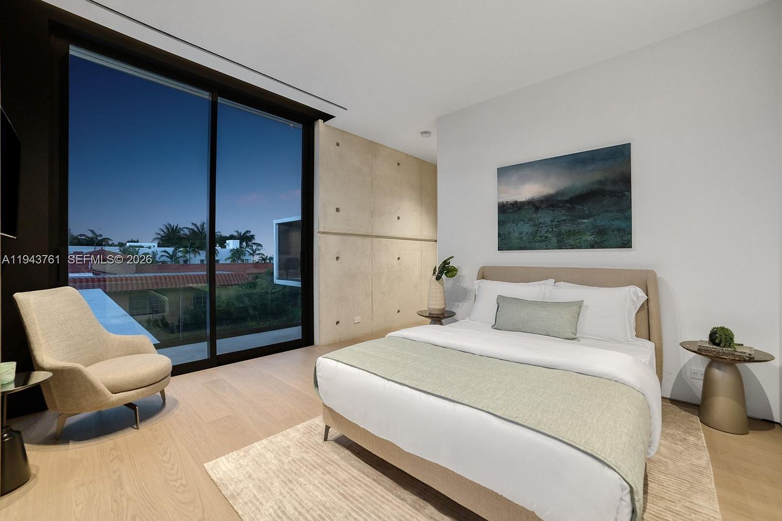 This modern bedroom features a minimalist aesthetic with light wood flooring and a prominent concrete accent wall. A plush bed with neutral bedding sits center stage, complemented by a contemporary armchair and side tables, while large glass sliding doors provide a seamless view of the outdoor pool area. The room is bathed in soft, ambient light, creating a serene and sophisticated atmosphere.