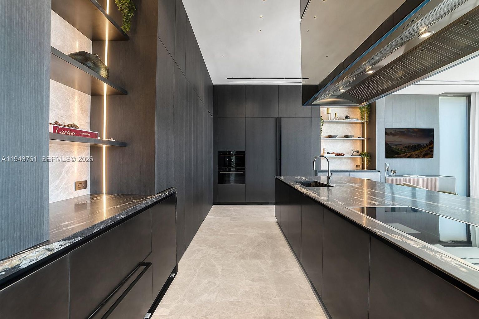 This sleek, contemporary kitchen features dark, minimalist cabinetry and a large central island topped with dark, veined stone. The space is accented by integrated linear lighting along open shelving and a professional-grade range hood suspended above the island. A neutral, light-toned stone floor provides a sharp contrast to the dark cabinetry, creating a sophisticated and high-end aesthetic.