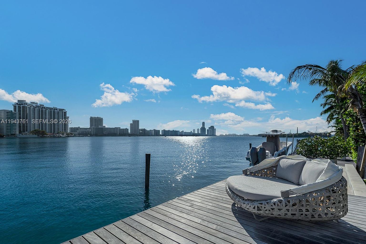 This image captures a serene waterfront deck featuring a modern, circular outdoor lounge chair with plush cushions. The wooden deck extends toward a calm, blue bay, with a boat docked nearby and a panoramic view of a distant city skyline under a bright, sunny sky. The perspective is from the deck looking out over the water, emphasizing the tranquil and luxurious lifestyle associated with waterfront living.