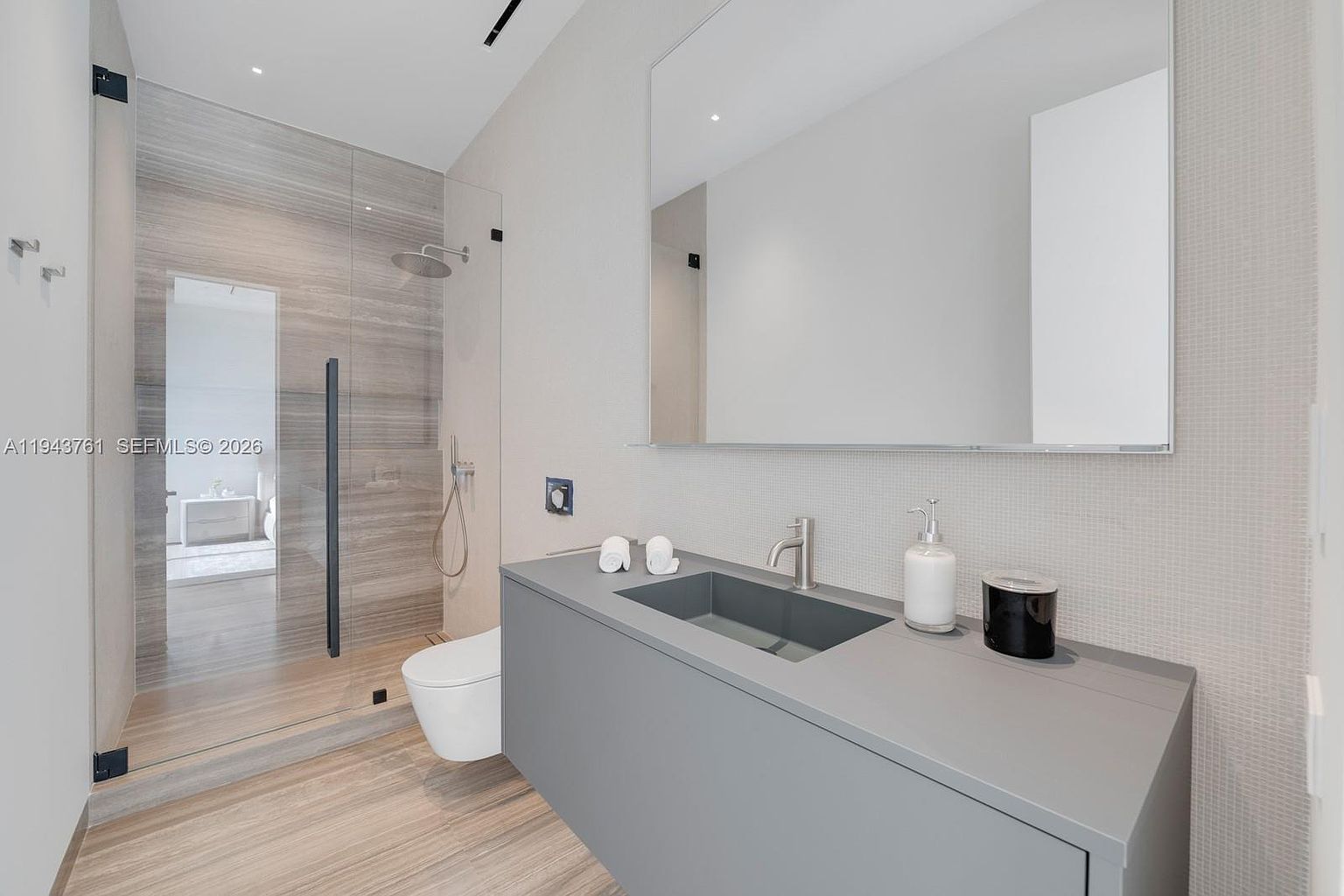 This modern bathroom features a sleek, minimalist vanity with a dark gray integrated sink and a large, frameless mirror. To the left, a walk-in shower is enclosed by glass panels and finished with light-toned, wood-look tile. The space is bright and contemporary, utilizing a neutral color palette and clean lines to create a sophisticated atmosphere.