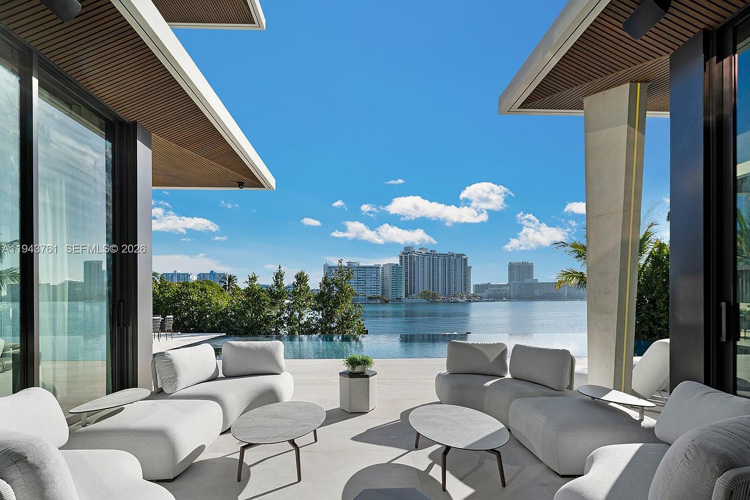 This luxurious outdoor living space features a covered patio furnished with plush, light-toned modular seating and modern oval coffee tables. The area seamlessly transitions to an infinity-edge pool that overlooks a serene bay and a distant city skyline. The open-air design, framed by architectural columns and expansive glass walls, creates a sophisticated atmosphere perfect for waterfront entertaining.