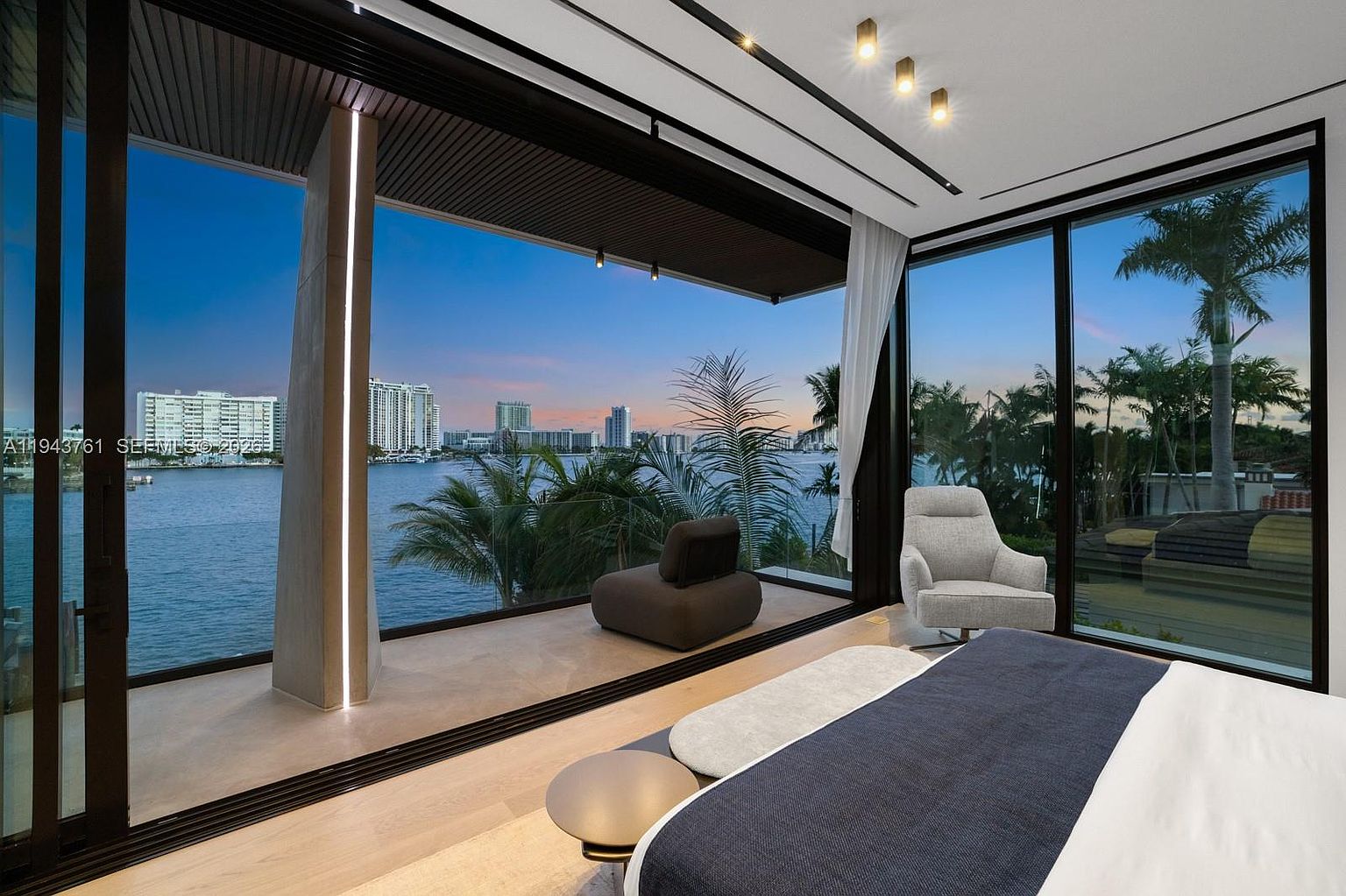 This luxurious primary bedroom features expansive floor-to-ceiling glass walls that provide an unobstructed, panoramic view of the waterfront and city skyline. The room is furnished with a plush, light-colored armchair and a dark, low-profile lounge chair on the private balcony, creating a serene retreat. The interior design is sleek and modern, characterized by light wood flooring, minimalist lighting fixtures, and a neutral color palette that emphasizes the dramatic outdoor scenery.