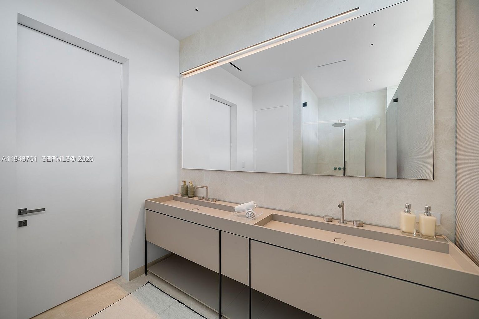 This modern, minimalist bathroom features a sleek, integrated double-sink vanity with a matte finish and clean lines. A large, frameless mirror spans the width of the vanity, reflecting the shower area and enhancing the sense of space in the room. The neutral color palette and contemporary fixtures create a sophisticated, spa-like atmosphere.