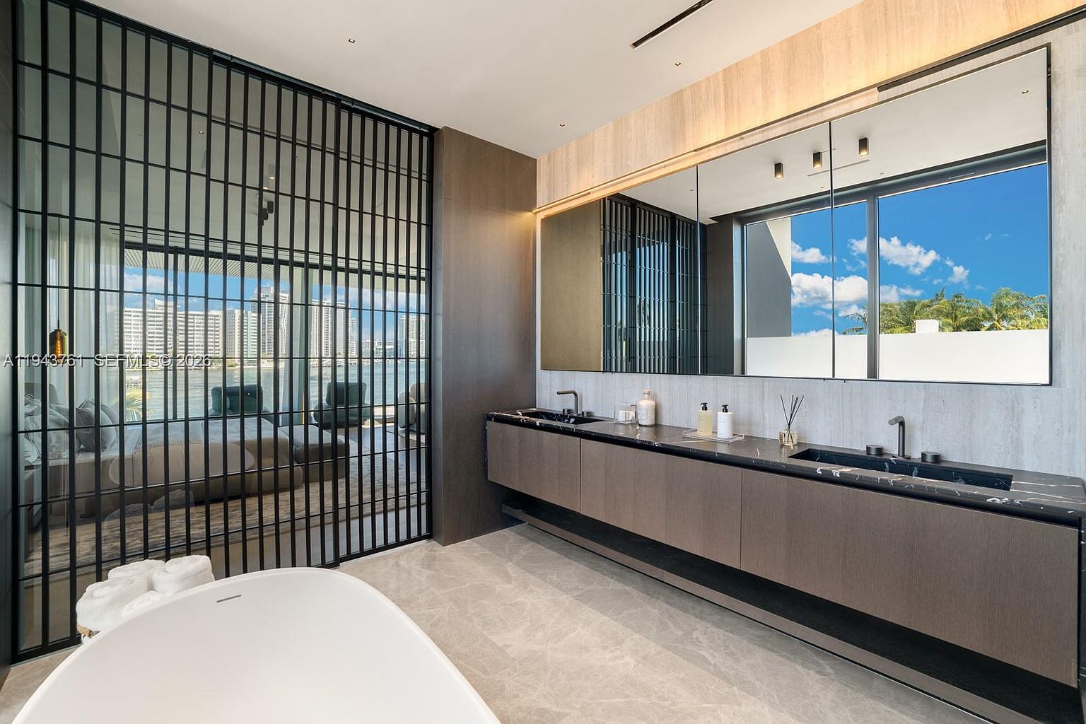 This luxurious primary bathroom features a sleek, floating double vanity with a dark stone countertop and integrated sinks, set against a backdrop of dark wood cabinetry. A large, freestanding soaking tub sits in the foreground, while a floor-to-ceiling metal screen provides a semi-transparent partition to the adjacent bedroom. The space is illuminated by a expansive mirror and recessed lighting, creating a sophisticated and modern atmosphere with a view of the city skyline.