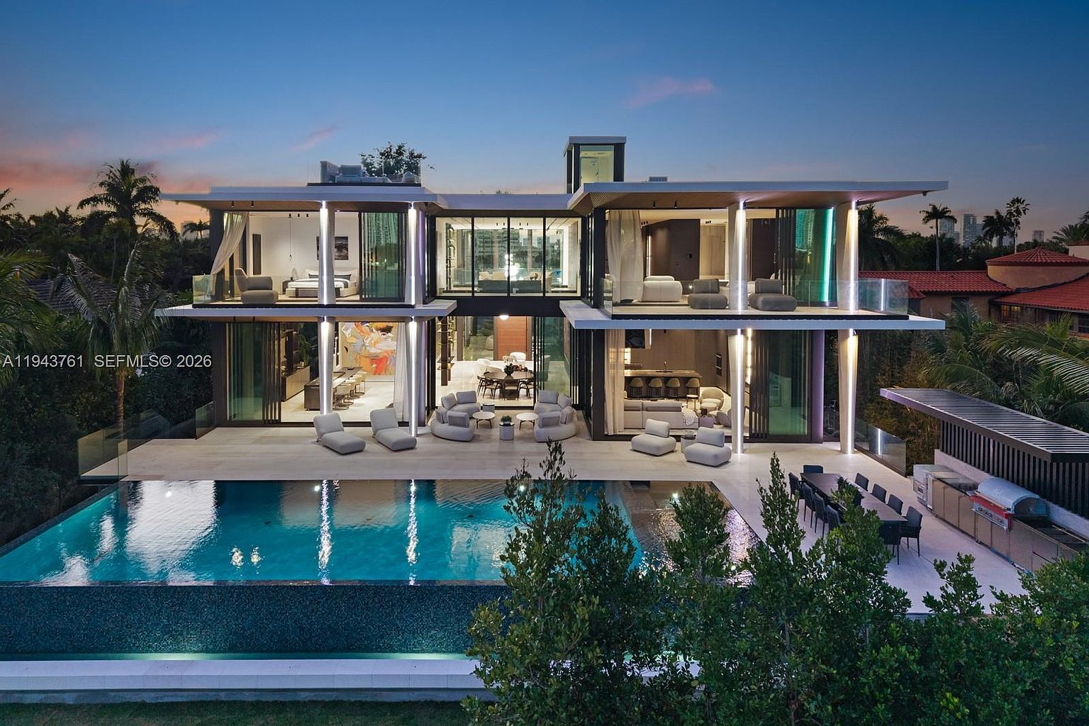 This modern, multi-level luxury residence is captured from an elevated rear perspective at dusk, showcasing a seamless indoor-outdoor living design. The home features extensive glass walls, multiple balconies, and a large infinity-edge pool that reflects the ambient lighting of the structure. A spacious patio area includes outdoor seating and a dedicated kitchen space, creating an ideal environment for high-end entertaining.
