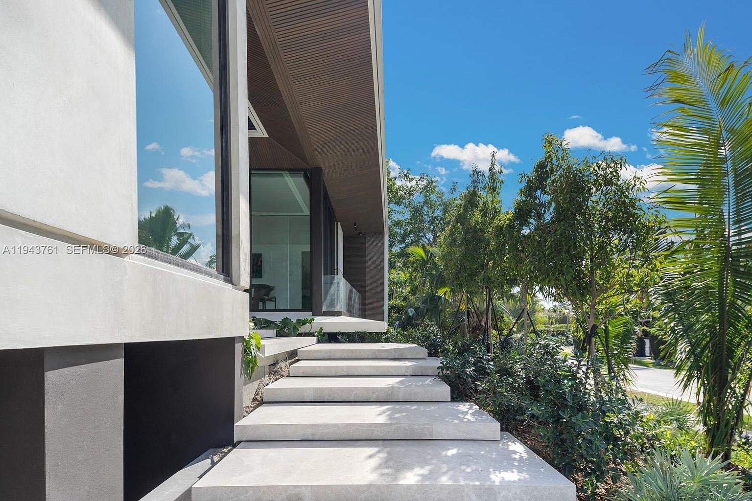 This modern entryway features a series of wide, floating concrete steps leading up to a sleek, contemporary home entrance. The architecture is defined by clean lines, large glass panels, and a warm-toned wood-slatted overhang, set against a backdrop of lush tropical landscaping. The perspective is low and angled, emphasizing the grand, geometric approach to the residence.