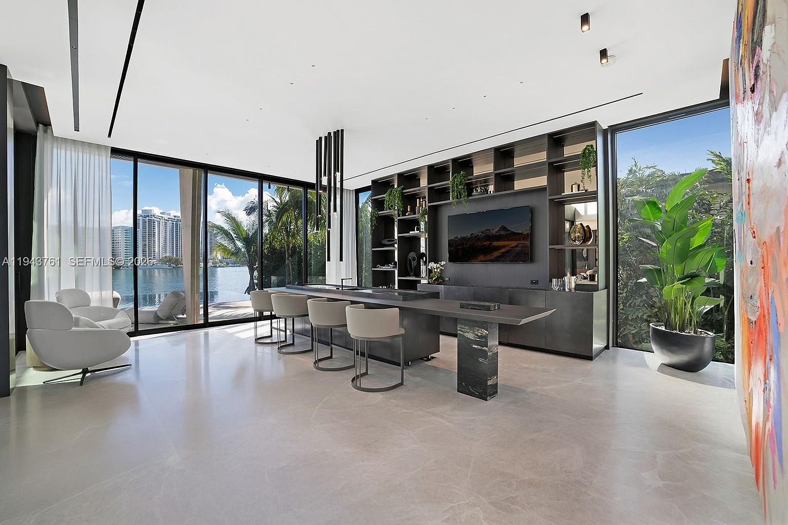 This modern, open-concept kitchen features a long, dark-toned island with integrated seating and a stone-clad base, serving as a central focal point. The space is defined by expansive floor-to-ceiling windows that provide a panoramic view of the waterfront, while a large, dark shelving unit with integrated lighting and a television anchors the back wall. The minimalist aesthetic is complemented by sleek, light-colored flooring and contemporary pendant lighting, creating a sophisticated and airy atmosphere.