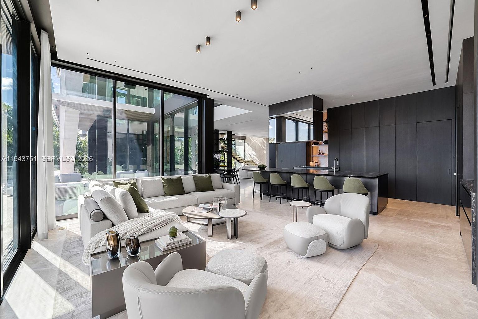 This expansive, open-concept living area features a sophisticated modern design with a neutral color palette and high-end finishes. The space includes a large sectional sofa, plush lounge chairs, and a sleek kitchen island with bar seating, all illuminated by recessed ceiling lights. Large floor-to-ceiling windows provide an abundance of natural light and a seamless connection to the outdoor patio area.