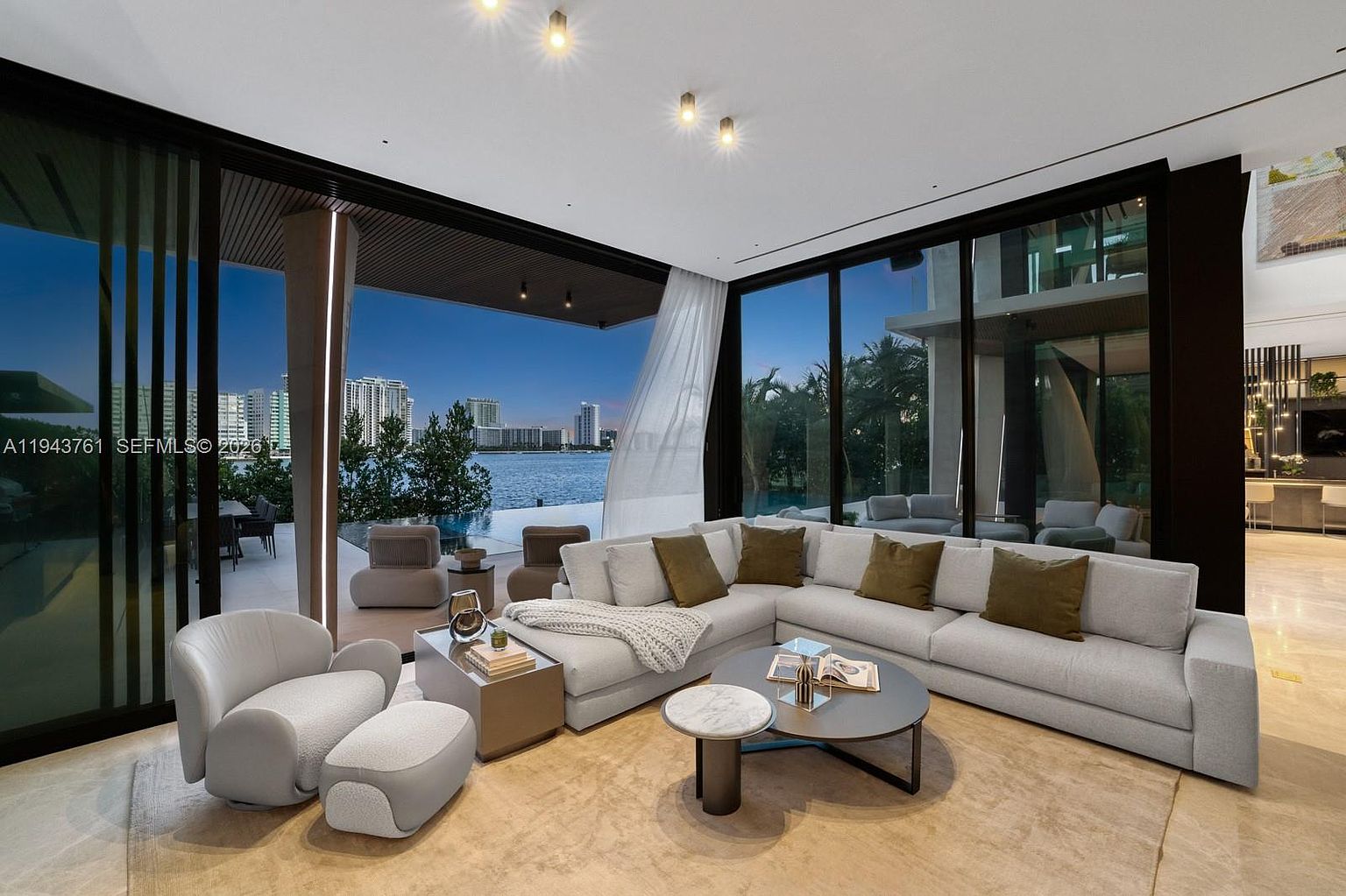 This sophisticated living room features an expansive open-concept layout with large glass walls that provide seamless indoor-outdoor flow to a waterfront terrace. The space is furnished with a plush, light-toned sectional sofa, a modern armchair with an ottoman, and a layered coffee table arrangement, all set atop a neutral area rug. The interior design emphasizes a contemporary aesthetic with recessed ceiling lighting and a clear view of the city skyline across the water.