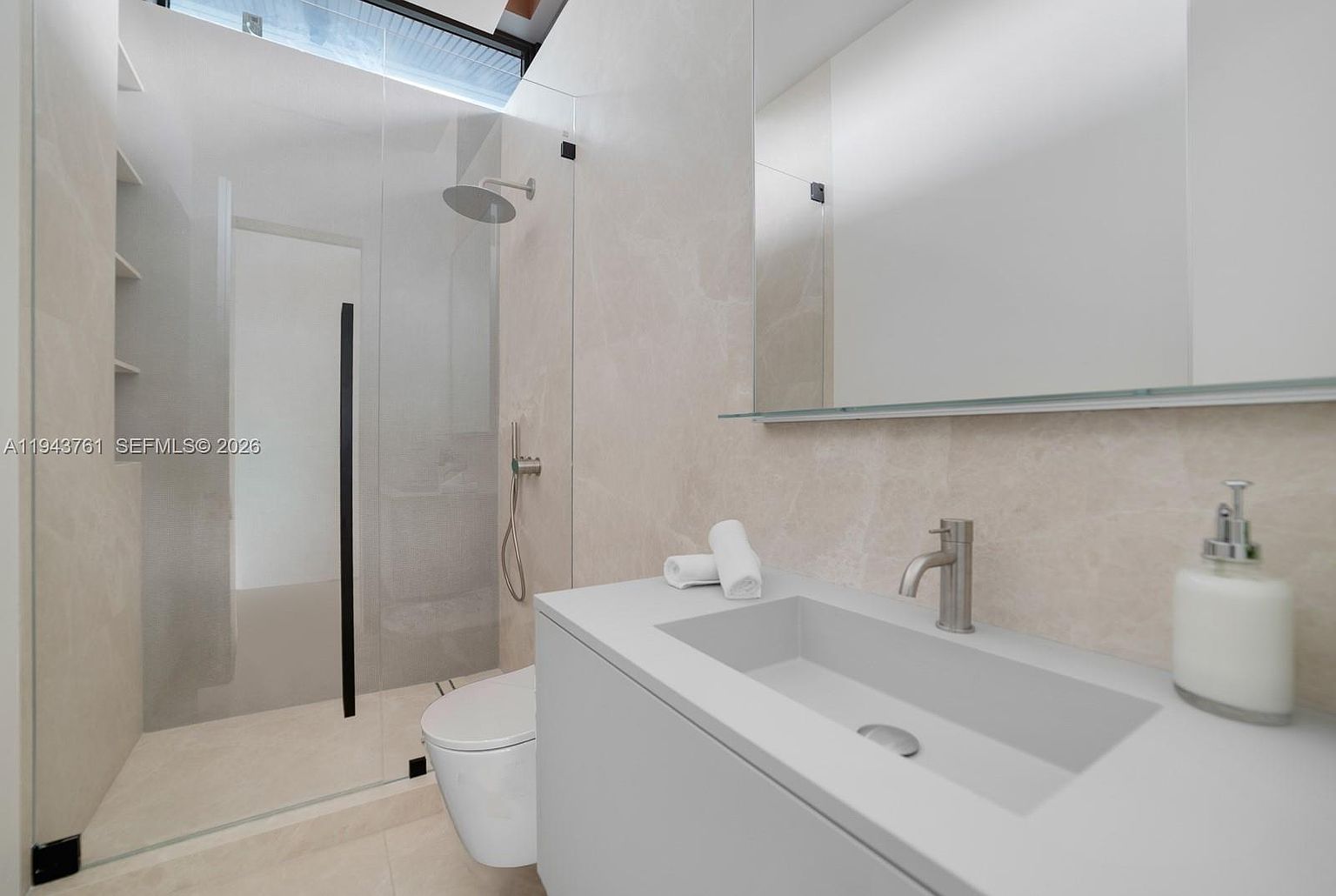 This modern bathroom features a sleek, minimalist vanity with a rectangular integrated sink and a tall, polished metal faucet. To the left, a glass-enclosed walk-in shower area is visible, complete with a rainfall showerhead and built-in shelving. The space is finished with light-toned, large-format wall tiles and a large, frameless mirror, creating a bright and airy atmosphere.