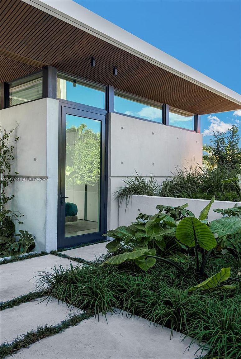 This modern entryway features a sleek, dark-framed glass door set against a textured concrete wall with visible architectural fasteners. A slatted wood overhang provides a warm contrast to the industrial aesthetic, while lush tropical landscaping and stone pavers create a welcoming path. The perspective is a low-angle shot that emphasizes the clean lines and contemporary design of the home's facade.