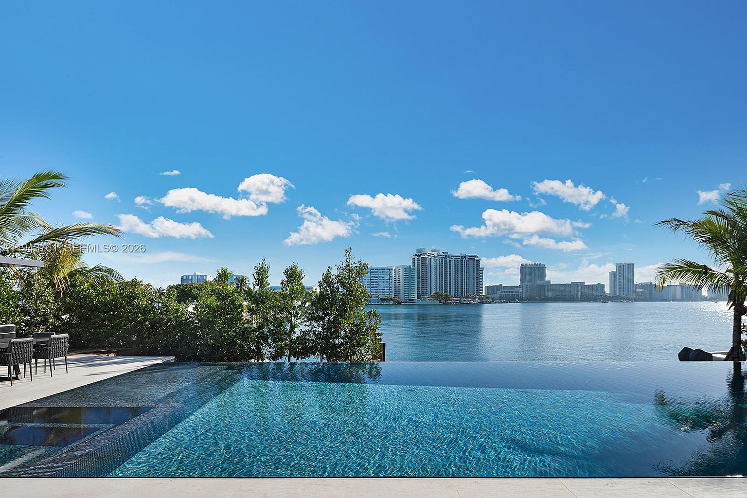 This image captures a stunning infinity-edge pool that seamlessly blends into the calm waters of the bay beyond. In the background, a skyline of high-rise buildings stands against a bright blue sky filled with scattered clouds, while lush green trees frame the left side of the scene. A small patio area with outdoor seating is visible on the left, creating a serene and luxurious atmosphere perfect for waterfront living.