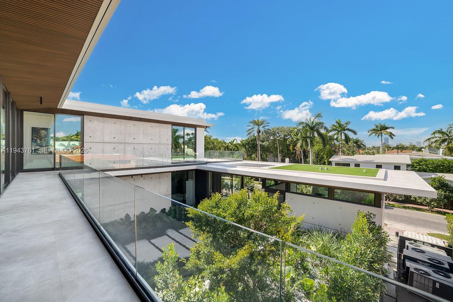 This image captures a modern, multi-level balcony with sleek glass railings overlooking a lush, landscaped courtyard. The architecture features clean lines, exposed concrete walls, and a flat roof design with a green roof section. The perspective is elevated, showcasing the seamless integration of indoor and outdoor living spaces in a contemporary residential setting.
