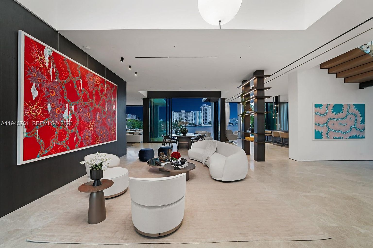This expansive, open-concept living area features a sophisticated modern aesthetic with light stone flooring and a striking, large-scale red abstract art piece mounted on a dark accent wall. The space is furnished with curved, contemporary white seating arranged around a tiered coffee table, while a tall, open-shelving room divider separates the living area from the dining space. Large glass sliding doors provide a seamless transition to the outdoors, revealing a nighttime view of a waterfront cityscape.