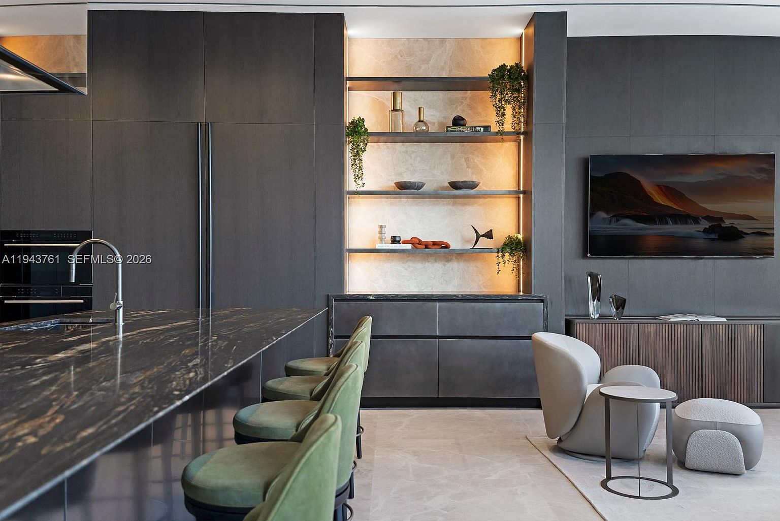 This modern kitchen features a large island with a dark, patterned stone countertop and a row of green upholstered bar stools. Behind the island, a recessed shelving unit with integrated lighting displays decorative objects against a light-toned stone backdrop, flanked by dark cabinetry. To the right, a comfortable seating area includes a curved armchair and a small side table, creating a seamless transition between the culinary and living spaces.