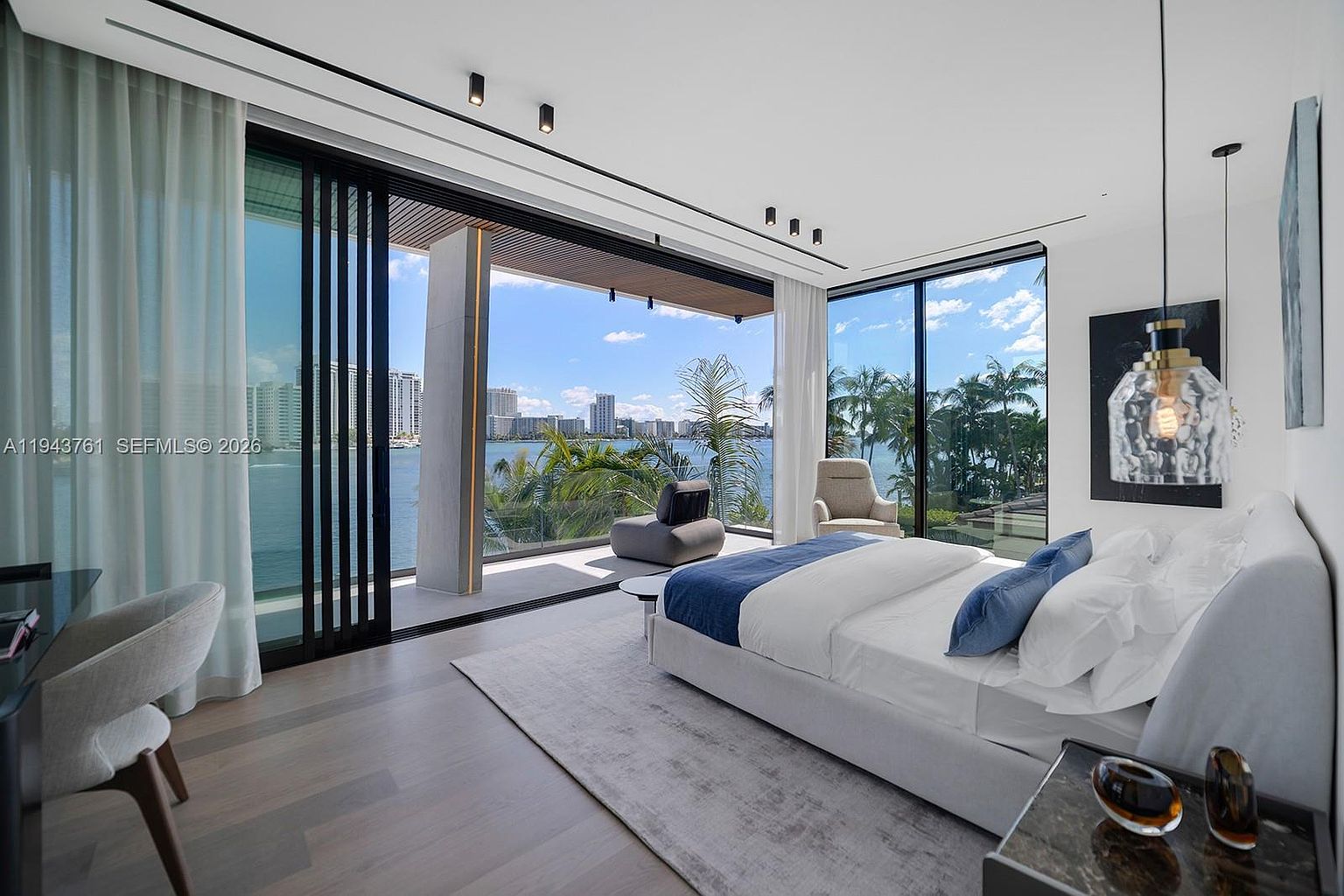 This luxurious primary bedroom features a minimalist aesthetic with a plush bed centered in the room, accented by blue pillows and a neutral area rug. Large floor-to-ceiling glass doors slide open to reveal a private balcony with panoramic water and city skyline views. The space is illuminated by recessed ceiling lights and a unique, textured glass pendant fixture, creating a serene and sophisticated atmosphere.