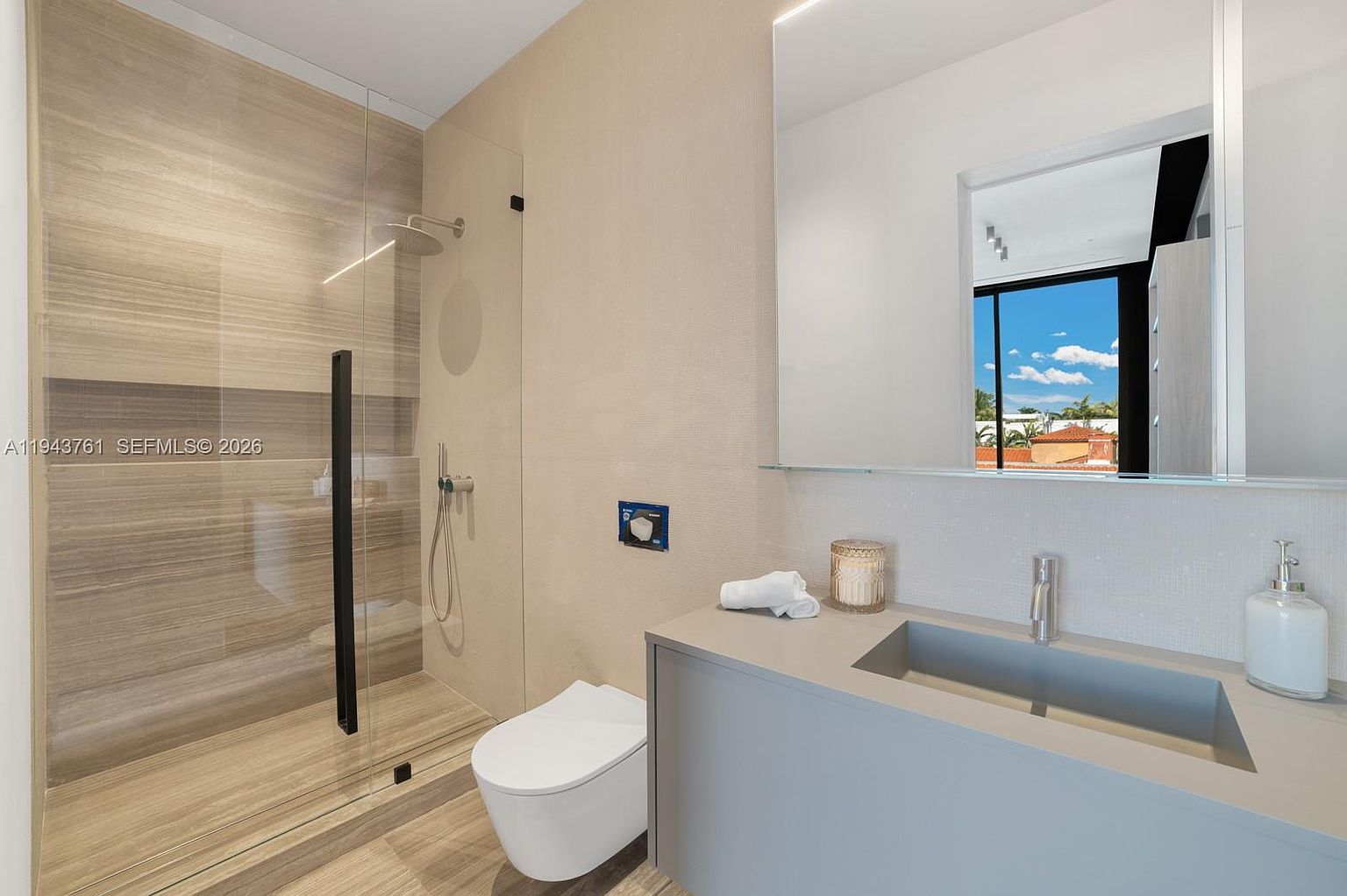 This modern bathroom features a sleek, minimalist design with a floating vanity and a walk-in shower enclosed by a glass panel. The space is finished with neutral-toned wall textures and wood-look tile flooring, creating a clean and contemporary aesthetic. A large, frameless mirror reflects the bright, airy atmosphere, while a wall-mounted toilet adds to the streamlined look.