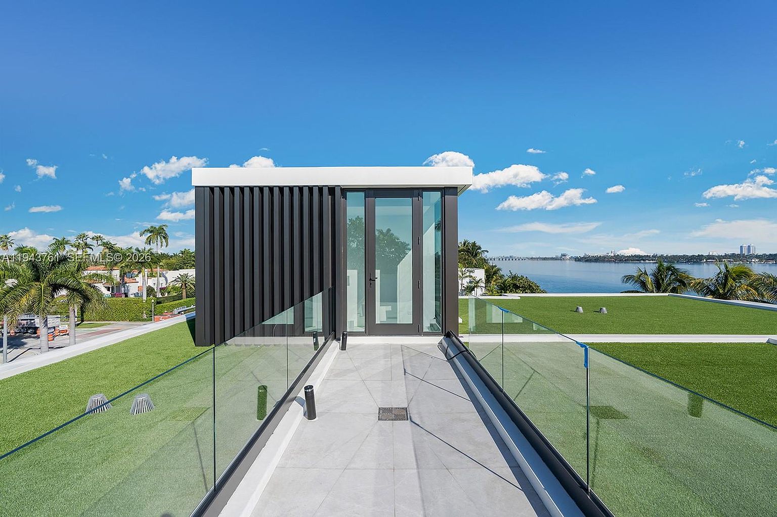 This rooftop terrace features a modern glass-enclosed walkway leading to a sleek, contemporary structure with dark vertical slats and large glass panels. The surrounding area is covered in vibrant green artificial turf, creating a clean and minimalist aesthetic. The perspective offers a wide, elevated view of the waterfront and distant horizon under a bright, clear blue sky.