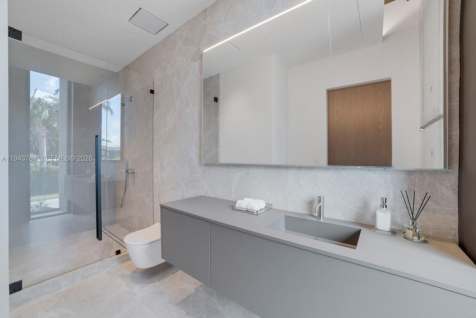 This modern bathroom features a sleek, wall-mounted gray vanity with an integrated sink and a large, minimalist mirror with integrated lighting. To the left, a glass-enclosed walk-in shower is visible, while the walls and floor are finished in large-format, light-toned stone tiles. The space exudes a clean, contemporary aesthetic with high-end finishes and a bright, airy atmosphere.
