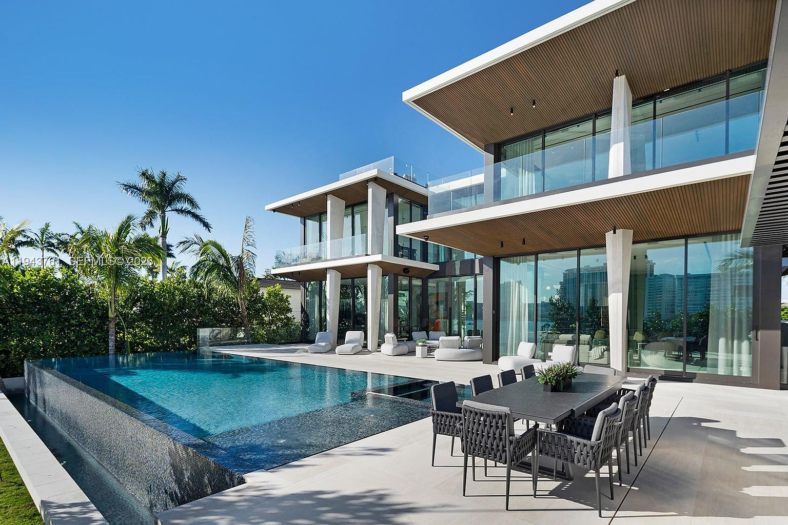 This modern luxury residence features a stunning infinity-edge pool that seamlessly blends with the outdoor patio area. The architecture showcases clean lines, expansive glass walls, and a covered dining terrace furnished with contemporary seating. Lush tropical landscaping and palm trees frame the view, creating a serene and sophisticated atmosphere perfect for high-end outdoor entertaining.