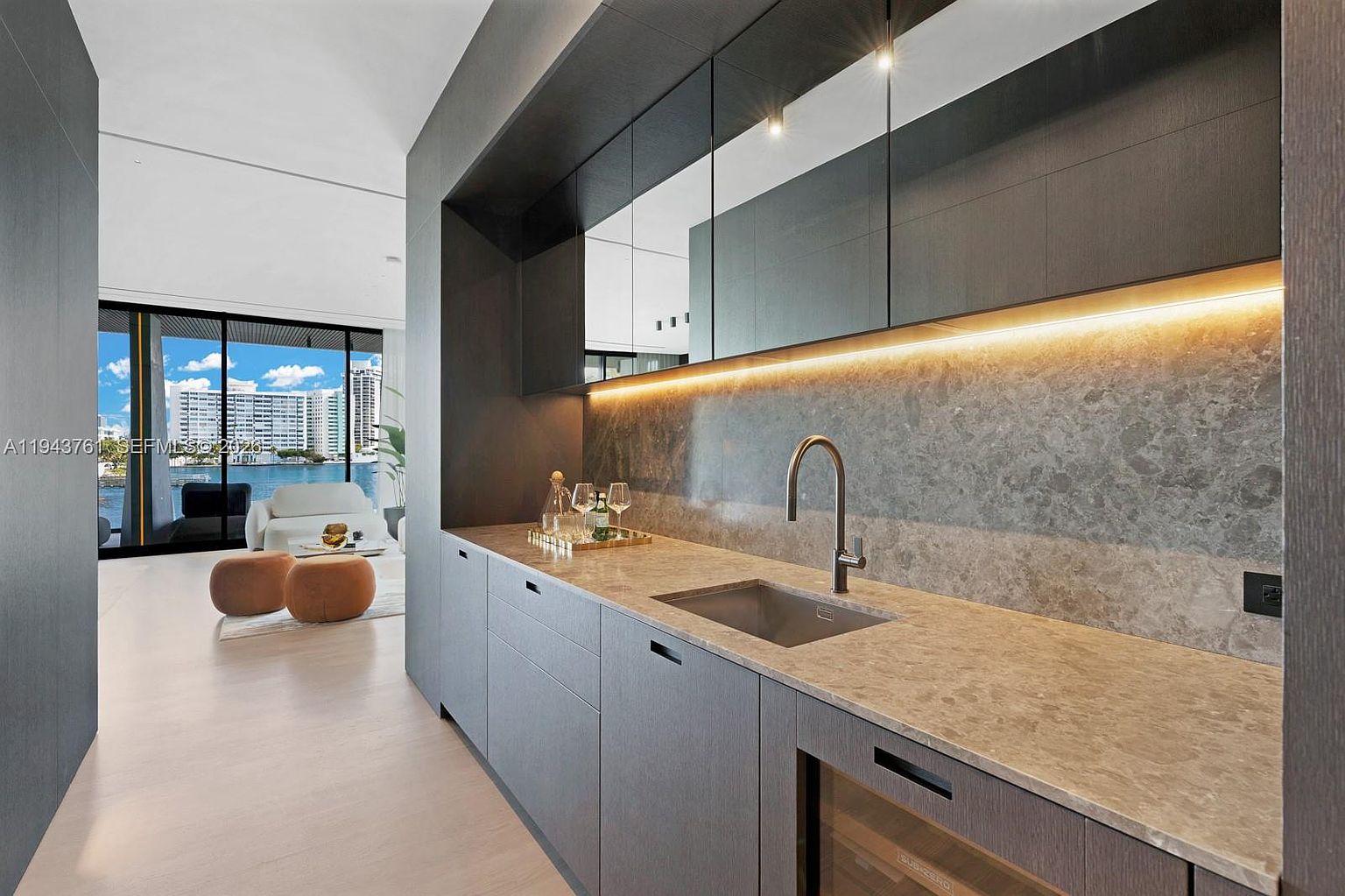 This sleek, modern wet bar or kitchenette features dark, textured cabinetry paired with a light-toned stone countertop and a matching backsplash. A stainless steel undermount sink with a gooseneck faucet is centered on the counter, while under-cabinet lighting highlights the workspace. The perspective looks past the bar into a bright living area with floor-to-ceiling glass doors that open to a waterfront view.