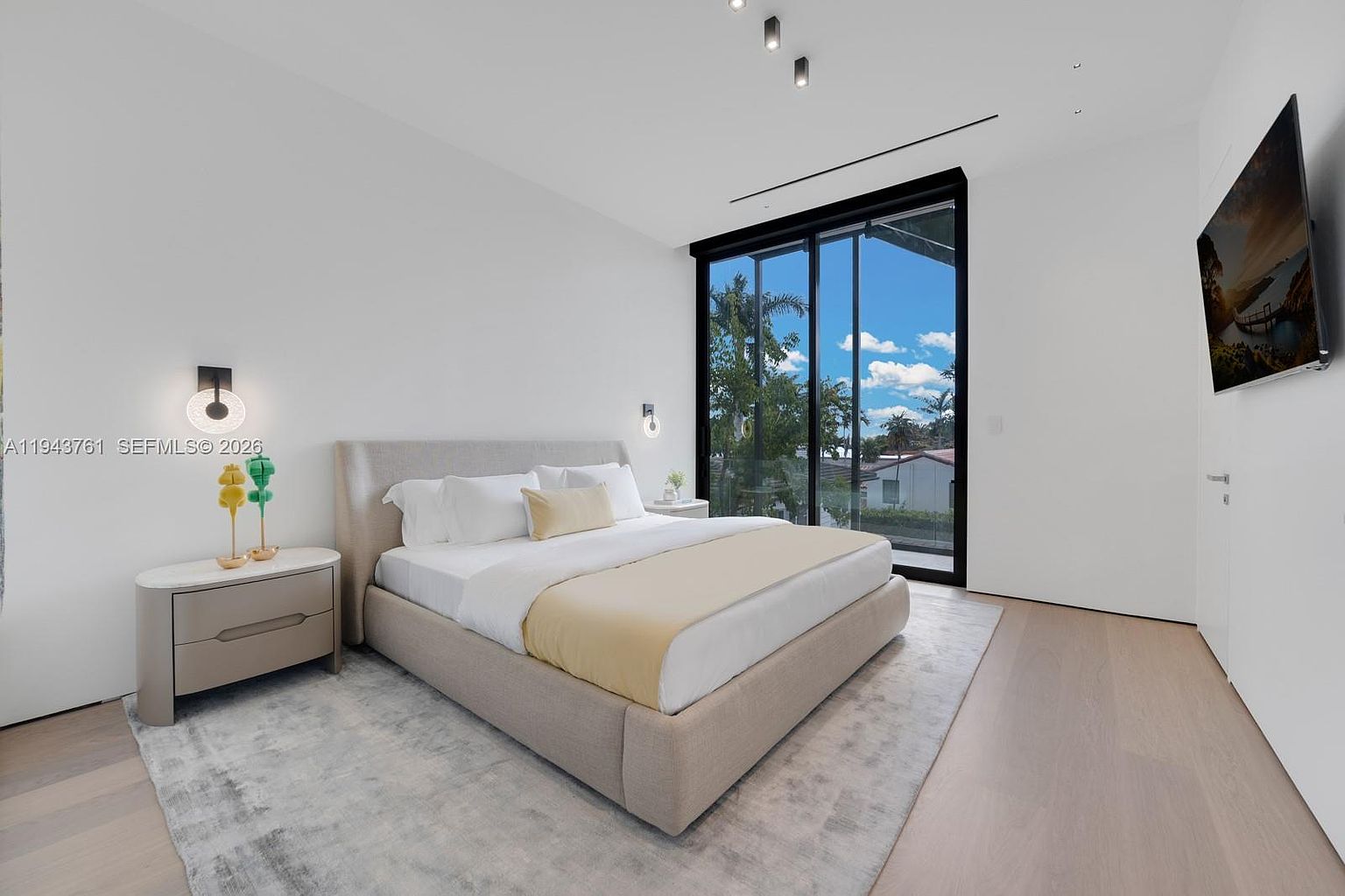 This modern primary bedroom features a neutral color palette with light wood flooring and a large, upholstered bed as the central focus. A wall of floor-to-ceiling glass sliding doors provides abundant natural light and a view of the outdoor surroundings, while a wall-mounted television is positioned opposite the bed. The minimalist design is complemented by a sleek bedside table with decorative lighting, creating a serene and contemporary atmosphere.