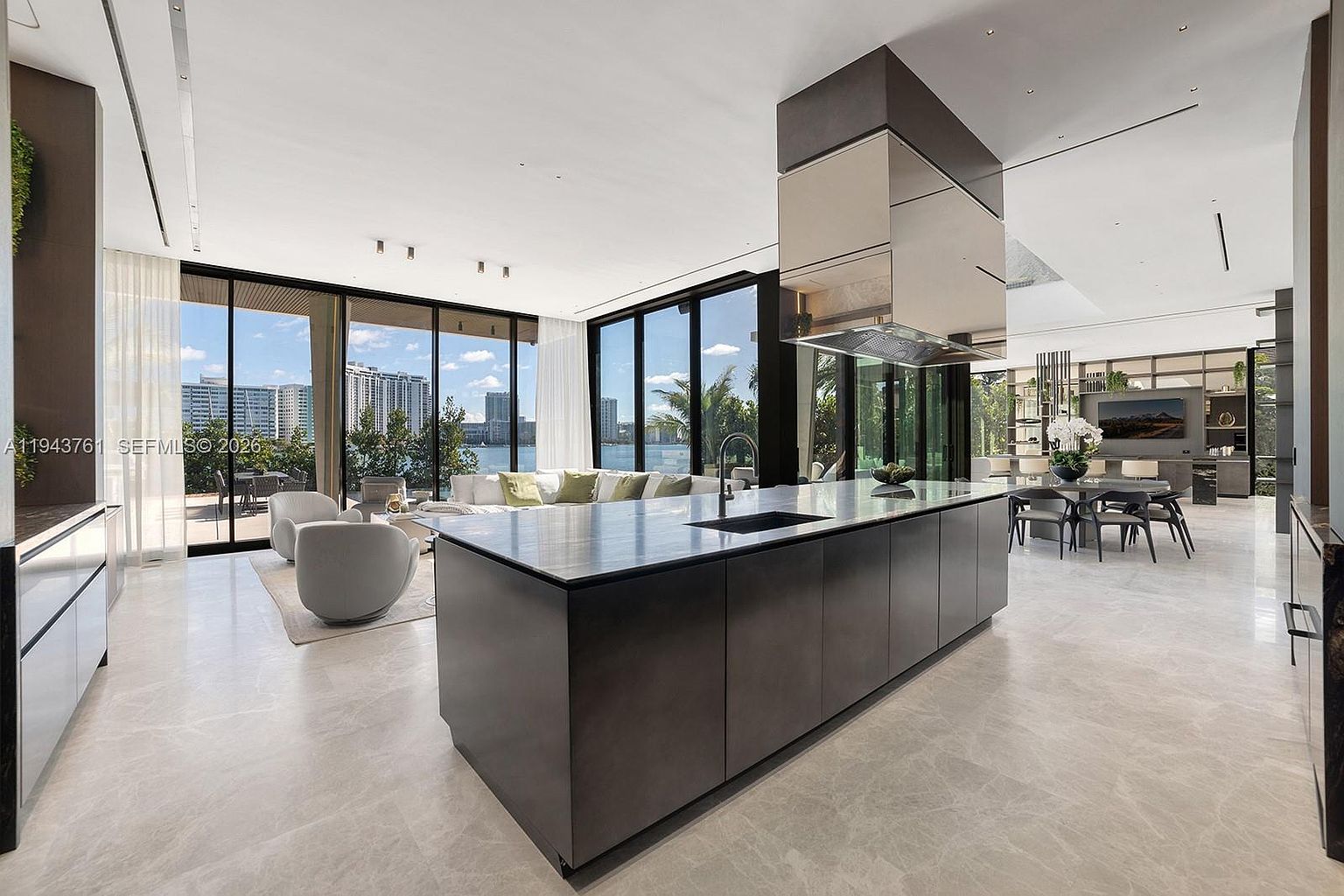This expansive, modern open-concept kitchen features a large, dark-toned central island with a stone countertop and an integrated sink. The space is defined by its sleek, minimalist aesthetic, highlighted by a prominent metallic range hood and light-colored stone flooring. Large windows provide an unobstructed view of the waterfront, seamlessly connecting the interior living area with the outdoor scenery.