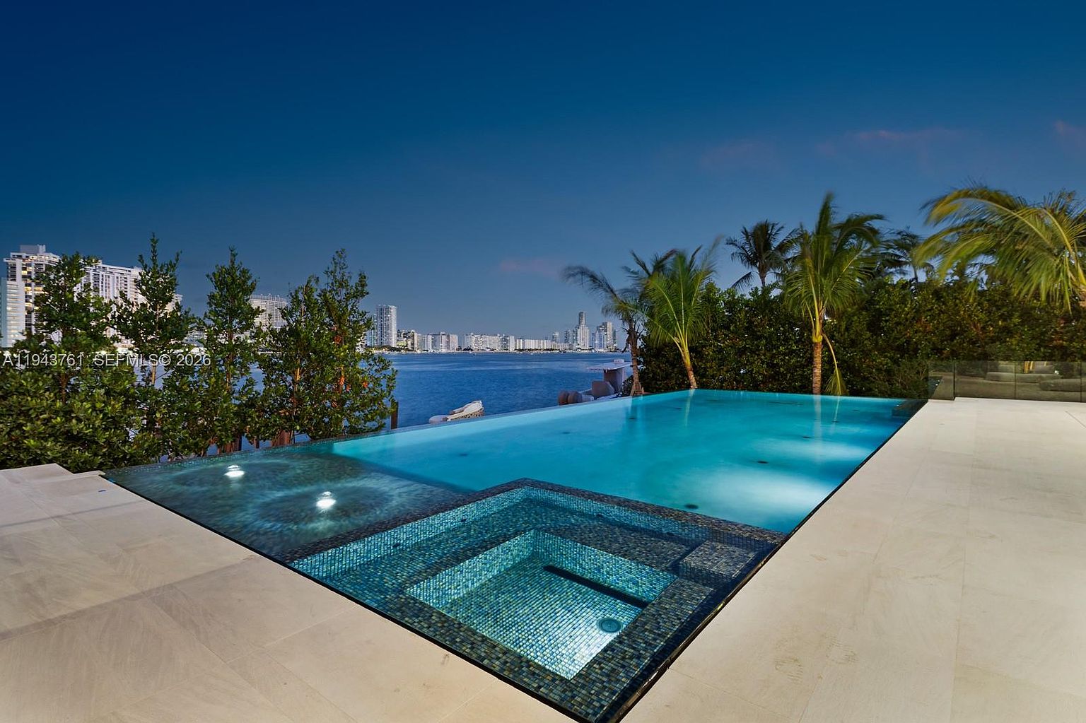 This stunning infinity-edge pool and integrated spa overlook a tranquil bay with a distant city skyline visible under a twilight sky. The pool deck is finished in light-toned stone, providing a clean, modern contrast to the deep blue water and the lush green trees lining the water's edge. The perspective is low and wide, emphasizing the seamless transition between the pool's edge and the horizon.