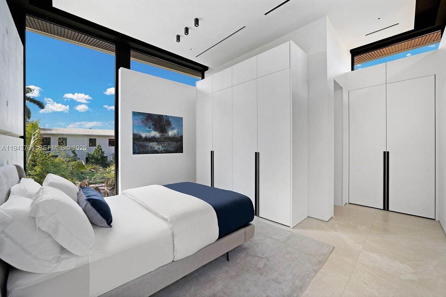 This modern bedroom features a minimalist aesthetic with a neutral color palette, light-toned tile flooring, and a large bed with crisp white linens. A wall of floor-to-ceiling windows provides abundant natural light and a view of the lush exterior greenery, while a large, built-in white wardrobe with dark vertical hardware offers ample storage. The room is accented by a piece of abstract art on the wall and recessed ceiling lighting, creating a clean and serene atmosphere.