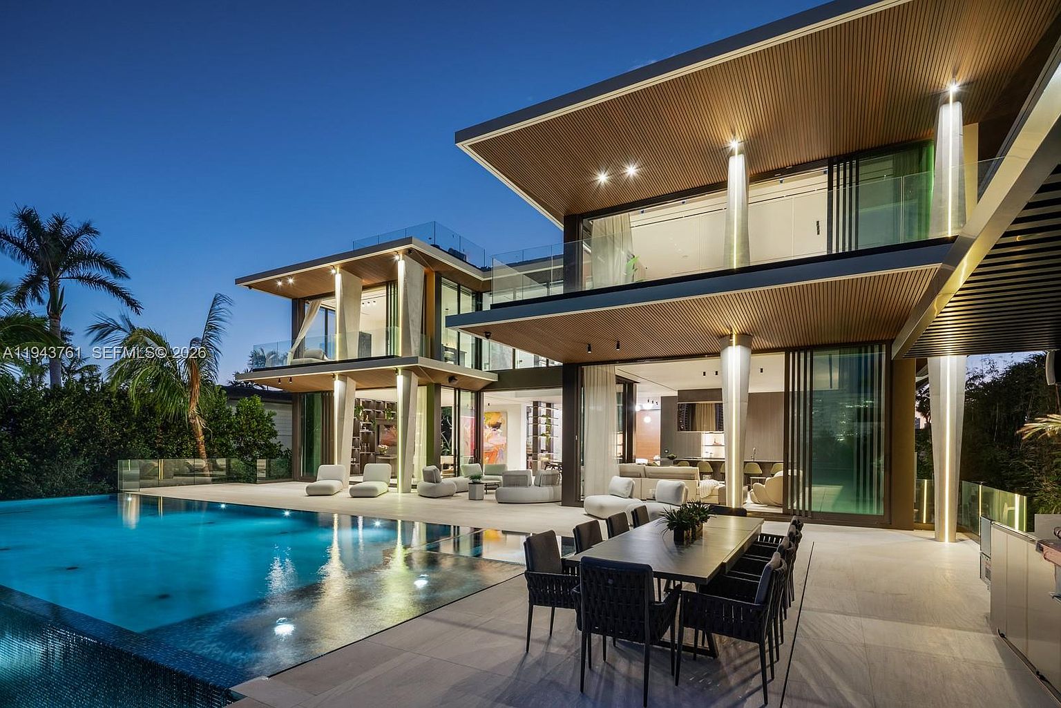 This stunning modern residence features a seamless indoor-outdoor transition, highlighted by a large infinity-edge pool and a spacious covered patio area. The architecture showcases clean lines, warm wood-toned soffits, and expansive glass walls that open to a sophisticated outdoor dining space. The scene is captured at twilight, emphasizing the dramatic lighting that illuminates the pool and the home's contemporary structural design.