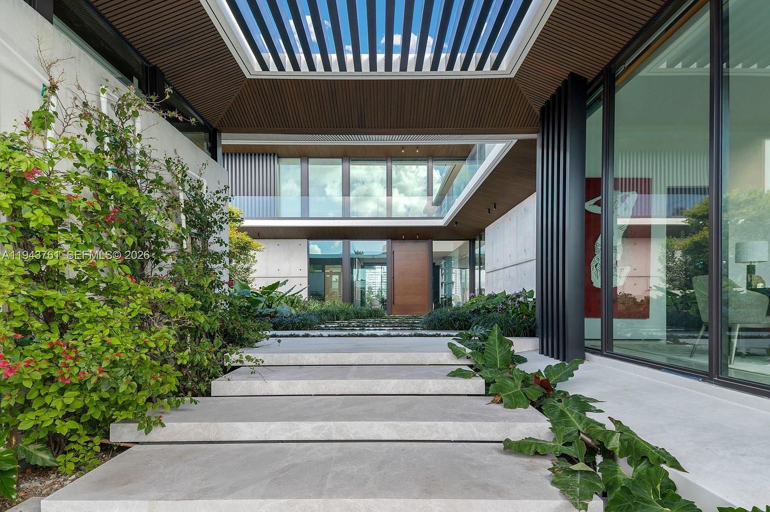 This striking modern entryway features a grand, tiered stone staircase leading to a prominent wooden front door. The architectural design incorporates a dramatic overhead slatted canopy, concrete walls, and expansive glass panels that offer a glimpse into the home's interior. Lush landscaping with vibrant greenery and flowering plants frames the entrance, creating a sophisticated and welcoming transition into the residence.