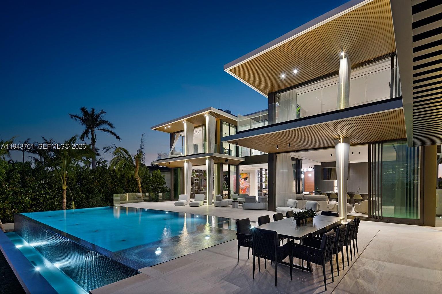 This modern luxury residence features a sprawling outdoor living area centered around a sleek, illuminated infinity-edge pool. The architectural design emphasizes indoor-outdoor flow with large glass walls, a covered patio dining space, and clean, contemporary lines. The twilight setting highlights the warm interior lighting and the sophisticated, resort-like atmosphere of the backyard.