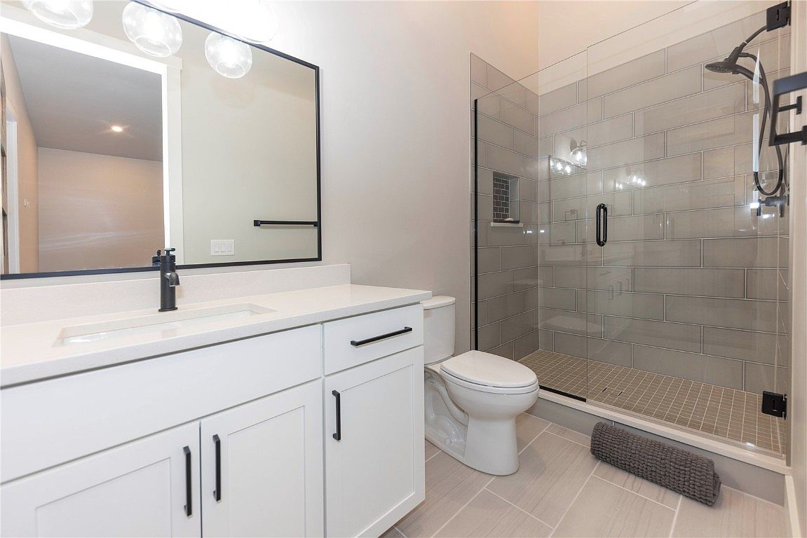This is a well-lit bathroom featuring a white vanity with black hardware, a modern black-framed mirror, and a toilet. The shower is enclosed with glass and has gray subway tile. The floor is tiled in a light gray tone, creating a clean and contemporary aesthetic.