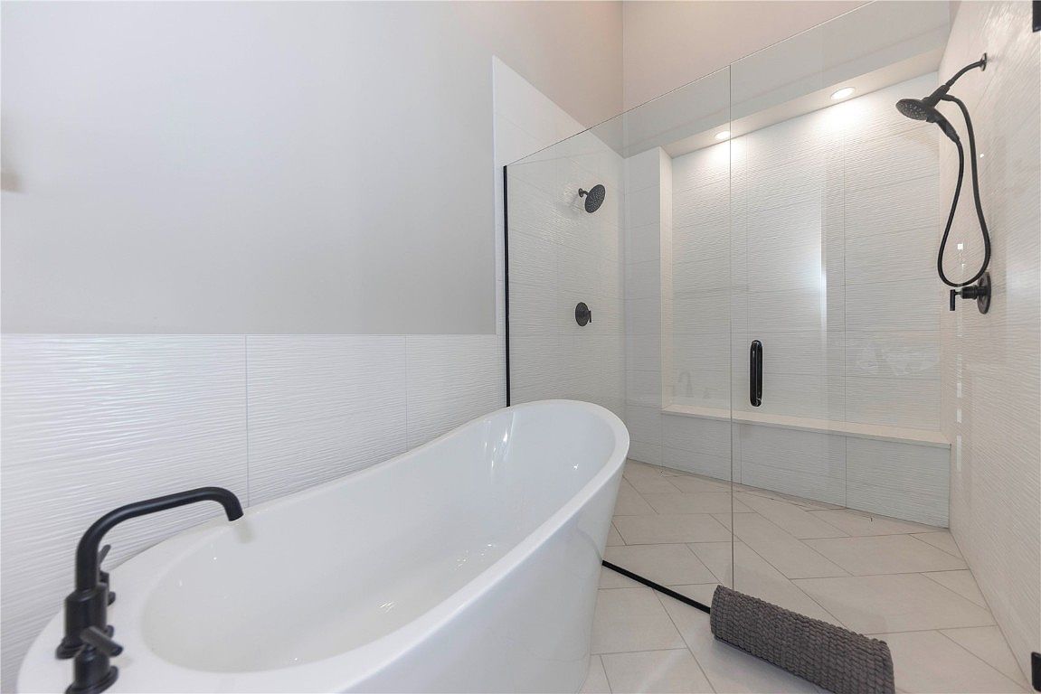 This is a bright and modern primary bathroom featuring a freestanding white bathtub with a black faucet. The walk-in shower has glass walls, textured white tiles, and black fixtures, including a rainfall showerhead. The floor is tiled in a light gray herringbone pattern, and a gray towel is neatly placed on the floor.