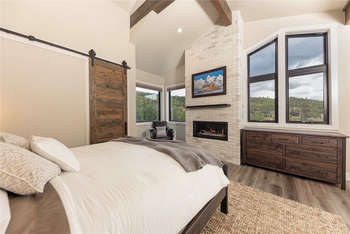 This is a primary bedroom featuring a bed with white linens and a gray throw blanket, a sliding barn door, and a brick fireplace with a mounted television. Large windows provide natural light and views of the surrounding landscape. A wooden dresser sits beneath the windows, complementing the hardwood flooring and creating a cozy, rustic-chic atmosphere.