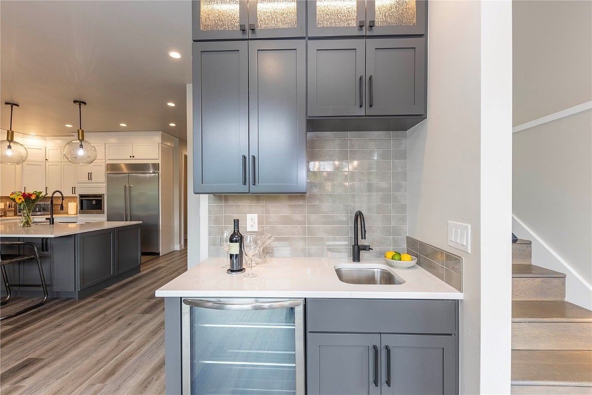 This interior shot showcases a modern kitchen with gray cabinetry and a white countertop. A wine fridge is built into the lower cabinets, and a small sink is present. The kitchen extends into the background with a large island and stainless steel appliances, creating a spacious and inviting atmosphere.