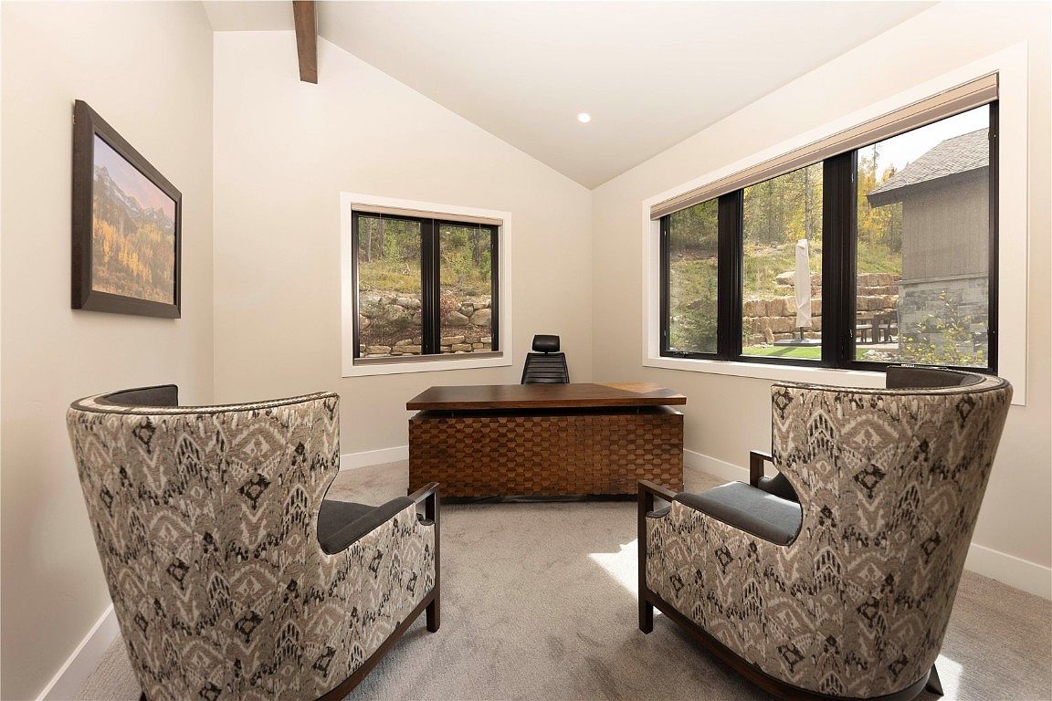 This is an interior shot of a modern office space featuring two patterned armchairs facing a wooden desk. The room has a high, angled ceiling and two windows that provide natural light and views of the outdoors. A framed picture hangs on the wall, adding a touch of personality to the space.