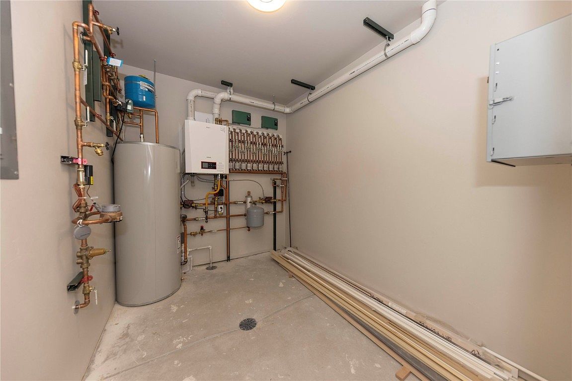 This is an interior shot of a basement utility room, featuring a water heater, pressure tank, and complex copper piping system along the left wall. A modern boiler and manifold system are installed on the back wall, with white PVC pipes running along the ceiling. The concrete floor is bare, and some trim pieces are stacked against the right wall, suggesting a functional, unfinished space.