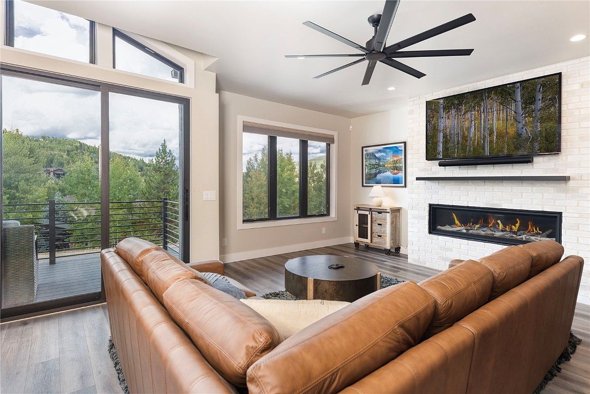 This is an interior shot of a modern living room featuring a large brown leather sectional sofa, a round metal coffee table, and a fireplace with a white brick surround. A large television is mounted above the fireplace, and a ceiling fan with multiple blades is visible. Sliding glass doors lead to an outdoor balcony with a view of trees and mountains, creating a bright and inviting space.
