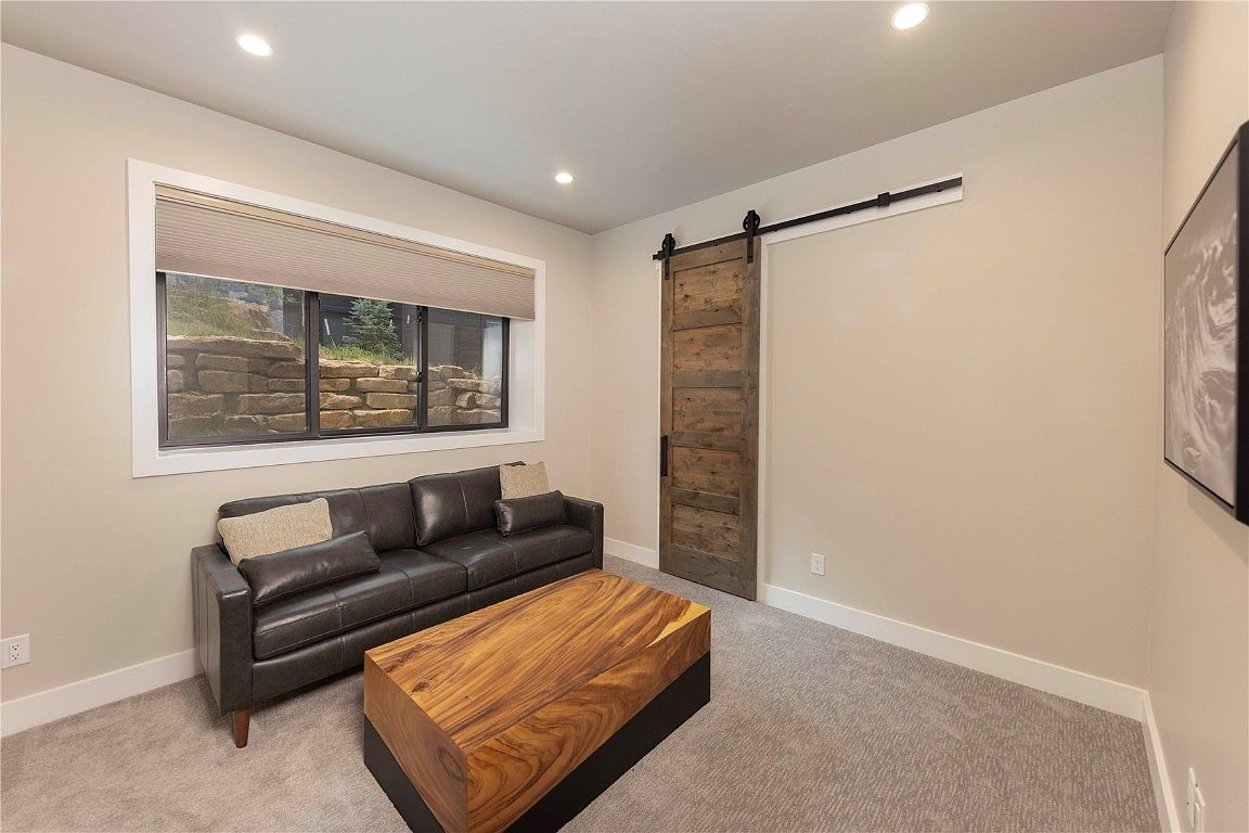 This is an interior shot of a cozy living room featuring a dark leather sofa with throw pillows, a unique wooden coffee table with a black base, and a sliding barn door. A window provides natural light, and the room is carpeted in a neutral tone. The overall impression is modern and comfortable.