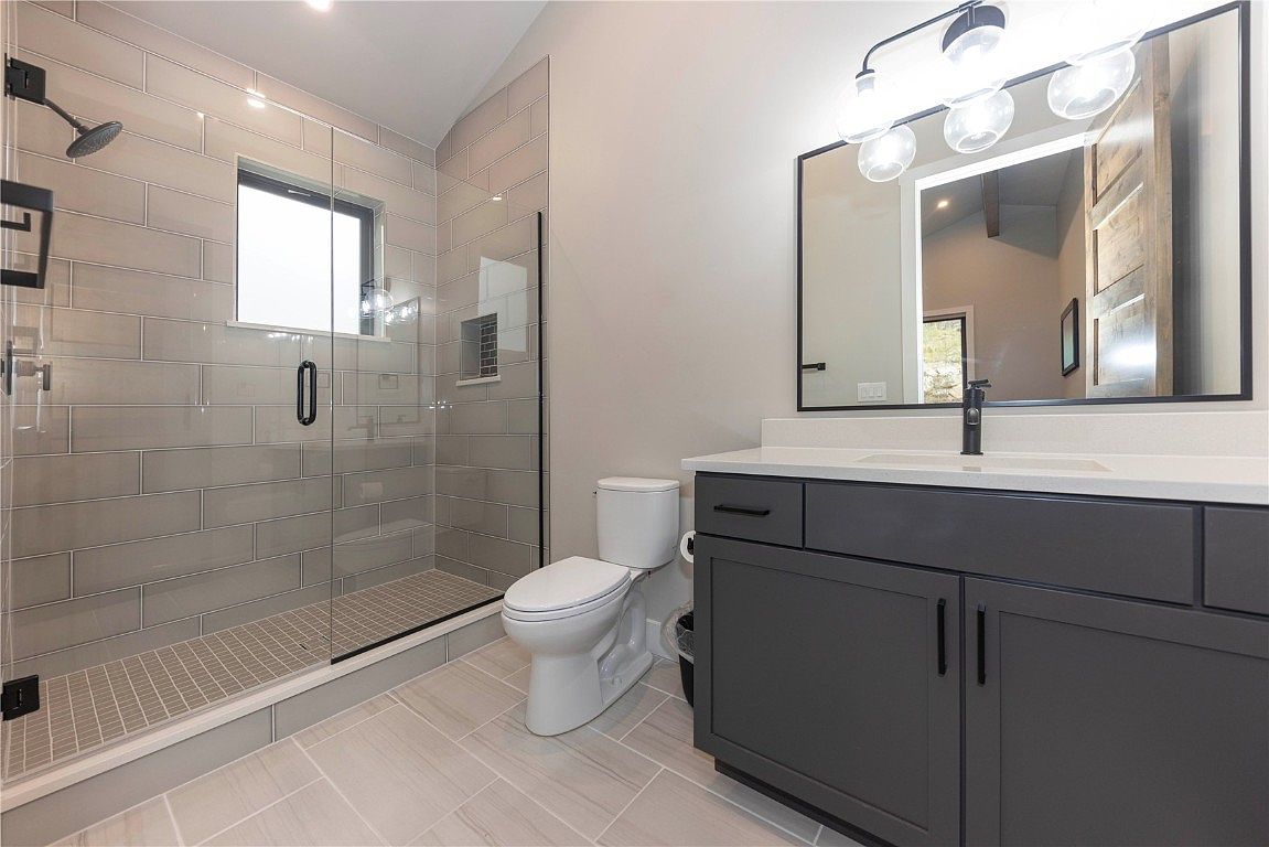 This is a modern bathroom featuring a glass-enclosed shower with gray subway tile and a grid-patterned floor. A gray vanity with a white countertop and a large mirror with globe lighting are also present. The overall design is sleek and contemporary, creating a clean and inviting space.
