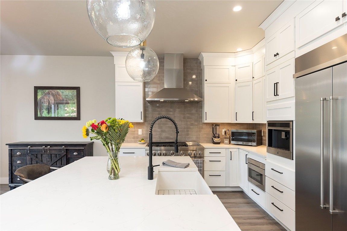This is a bright and modern kitchen featuring white cabinetry, stainless steel appliances, and a large island with a white countertop. The kitchen is well-lit with pendant lighting and recessed lights, and the backsplash is a neutral tile. The perspective is from the island, showcasing the sink, faucet, and the overall layout of the kitchen.