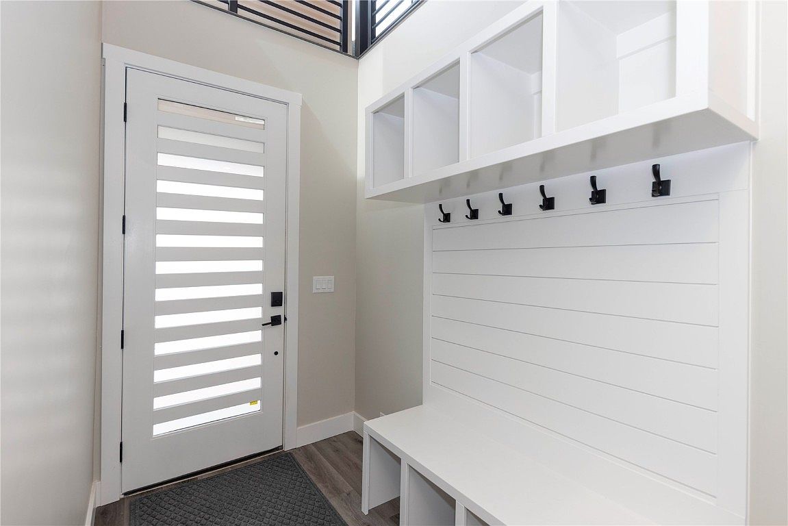 This interior shot showcases a modern entryway with a white door featuring horizontal window slats. A built-in storage unit with open shelving and coat hooks sits adjacent to the door, complemented by a bench with additional storage underneath. The space is well-lit and features neutral wall colors and wood-look flooring, creating a clean and inviting atmosphere.
