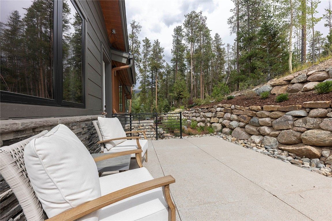 This image showcases a serene patio area adjacent to a modern home. The patio features comfortable outdoor seating with white cushions, set against a backdrop of a stone retaining wall and lush trees. The space offers a relaxing outdoor experience, perfect for enjoying the natural surroundings.